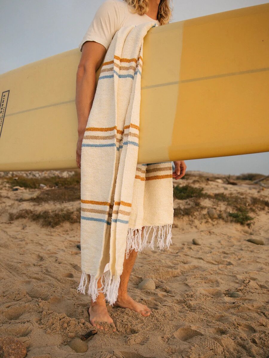 Person standing on a sandy beach holding a yellow surfboard, with a striped towel draped over their shoulder.