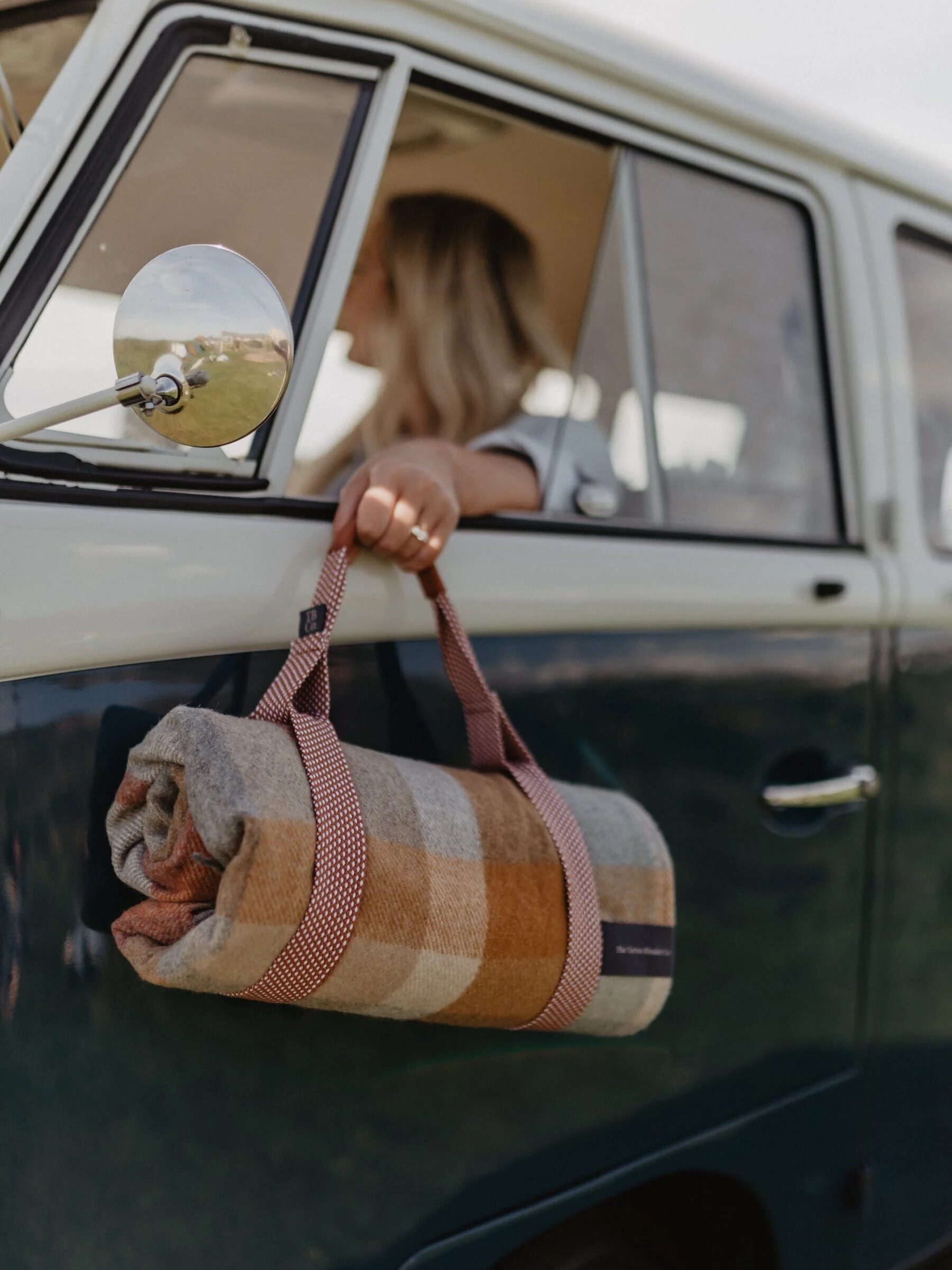 A person inside a vintage vehicle holds out a rolled plaid blanket through the window.