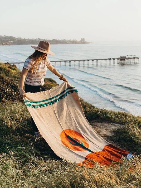 A person in a hat spreads out a colorful blanket on a grassy hill overlooking the ocean and a pier.