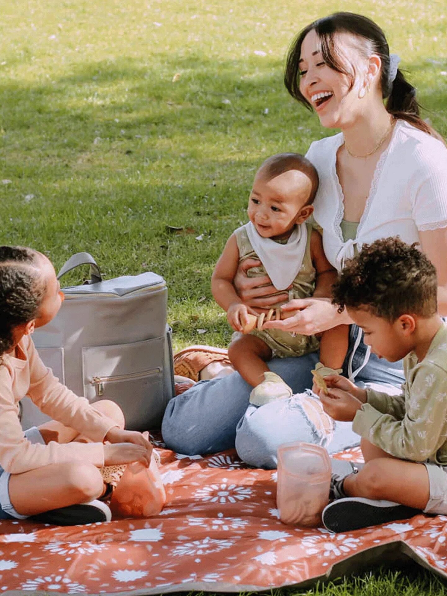 A woman sits on a picnic blanket with three children, two seated and one on her lap. They are enjoying a picnic outdoors on a sunny day.