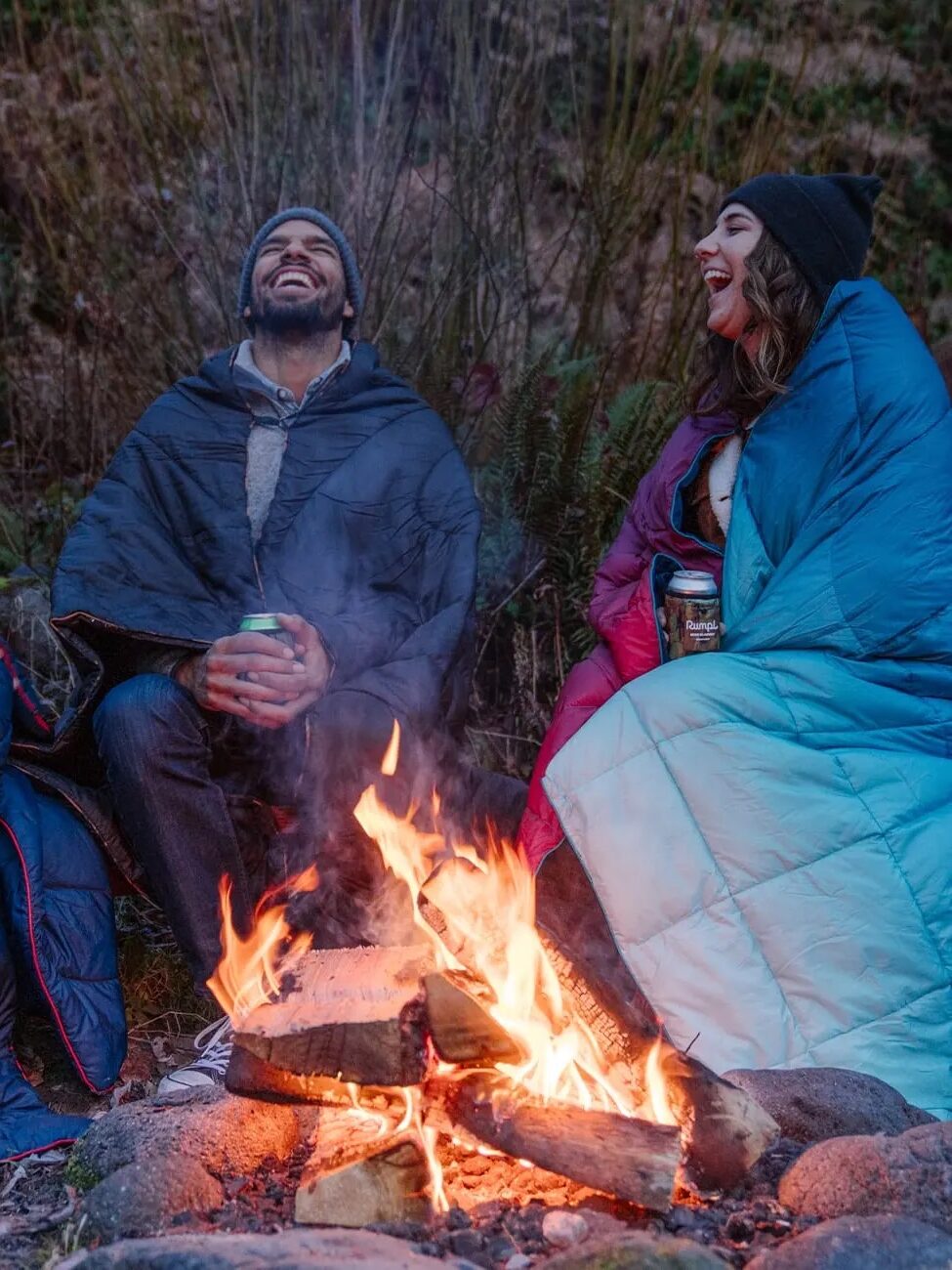 Three people wearing winter clothing and wrapped in blankets sit around a campfire, laughing and holding canned drinks.