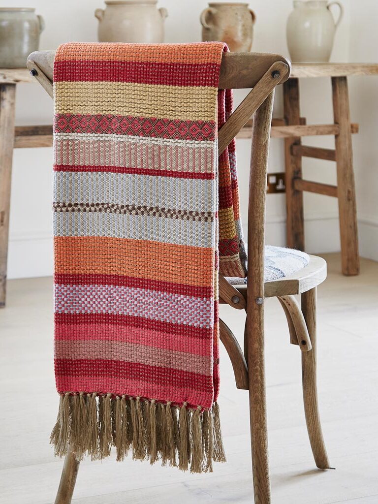 A multi-colored, striped blanket with fringed edges is draped over a wooden chair, placed in front of a rustic table with ceramic pots.