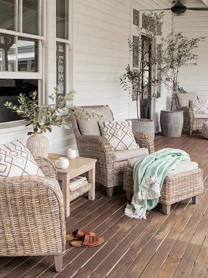 A porch with woven chairs, a small table, potted plants, and a green blanket on an ottoman.