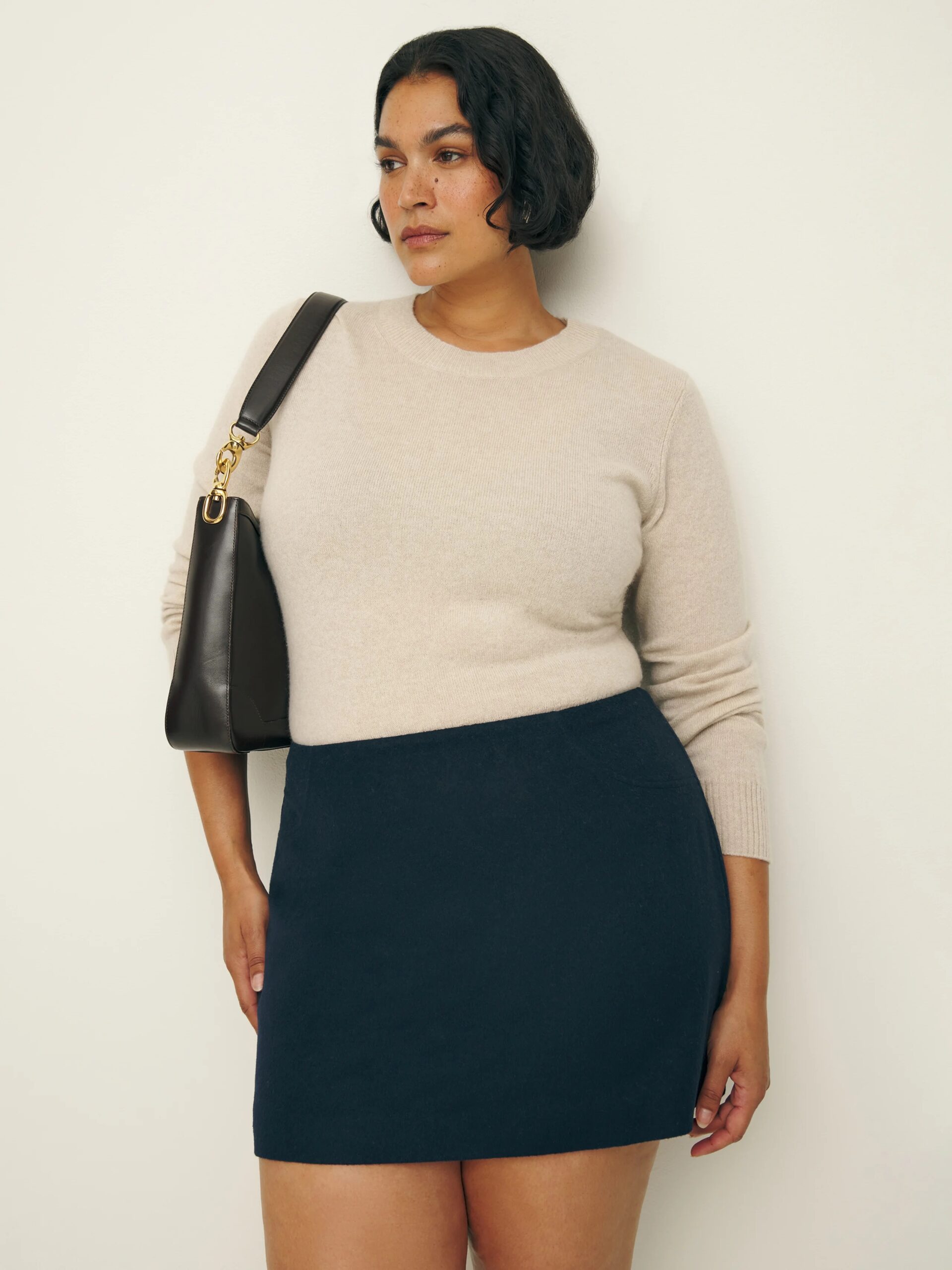 A plus size model wearing a beige long sleeve sweater a black miniskirt from Reformation. 