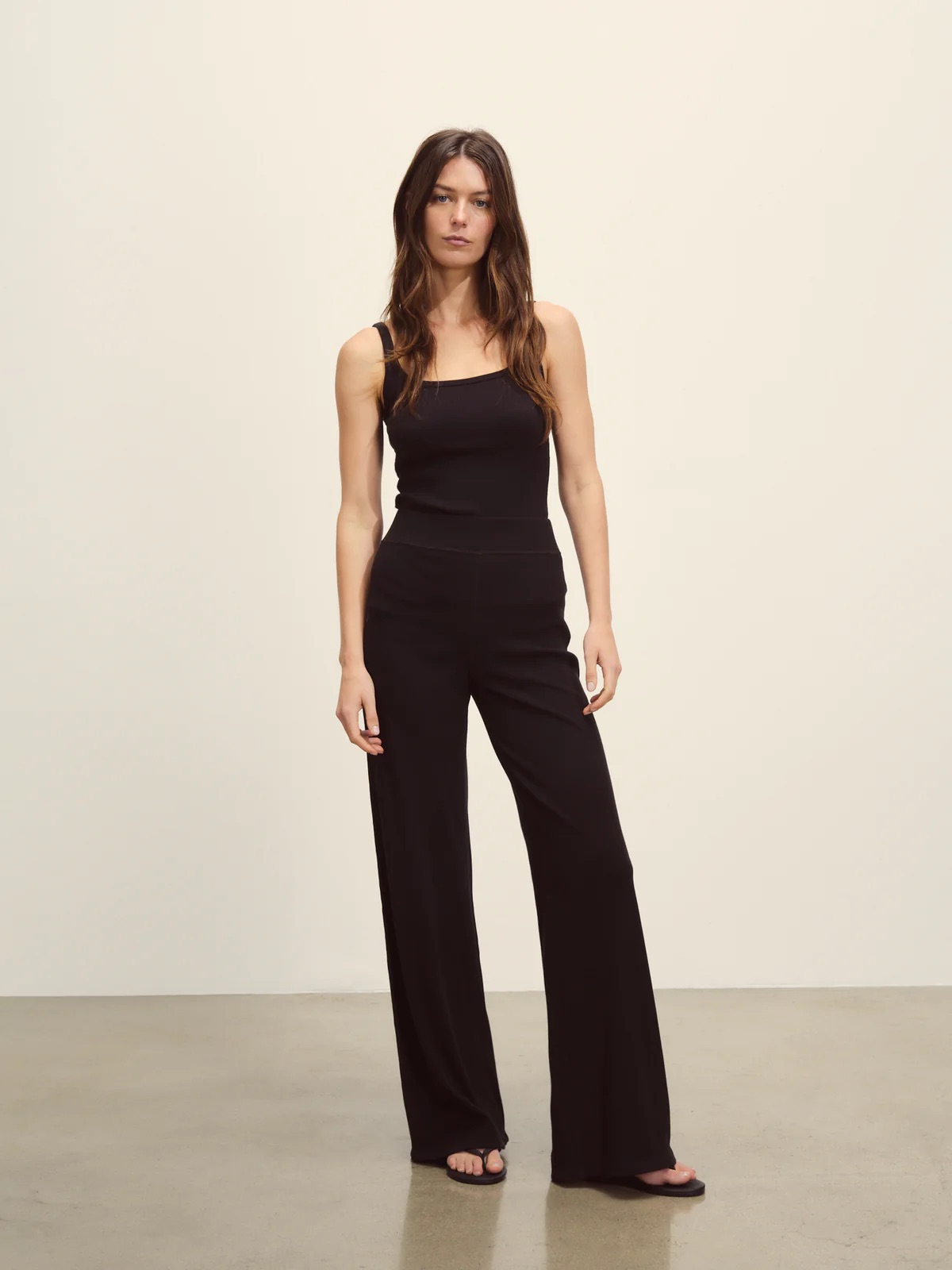 A woman stands on a plain background wearing a sleeveless black jumpsuit with wide legs and black sandals.