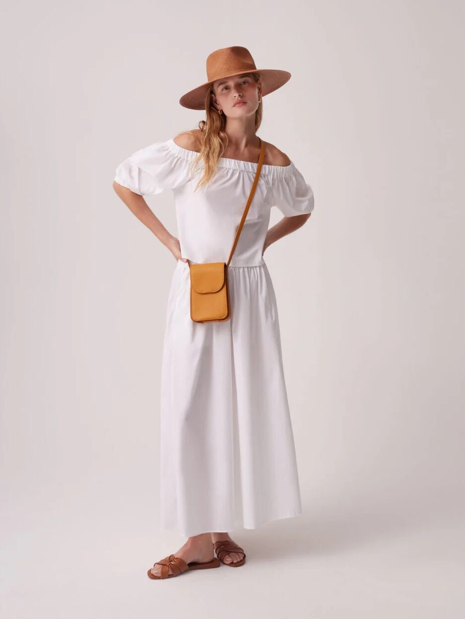 A woman wearing a white off-the-shoulder dress, brown hat, brown sandals, and an orange crossbody bag stands against a plain background.