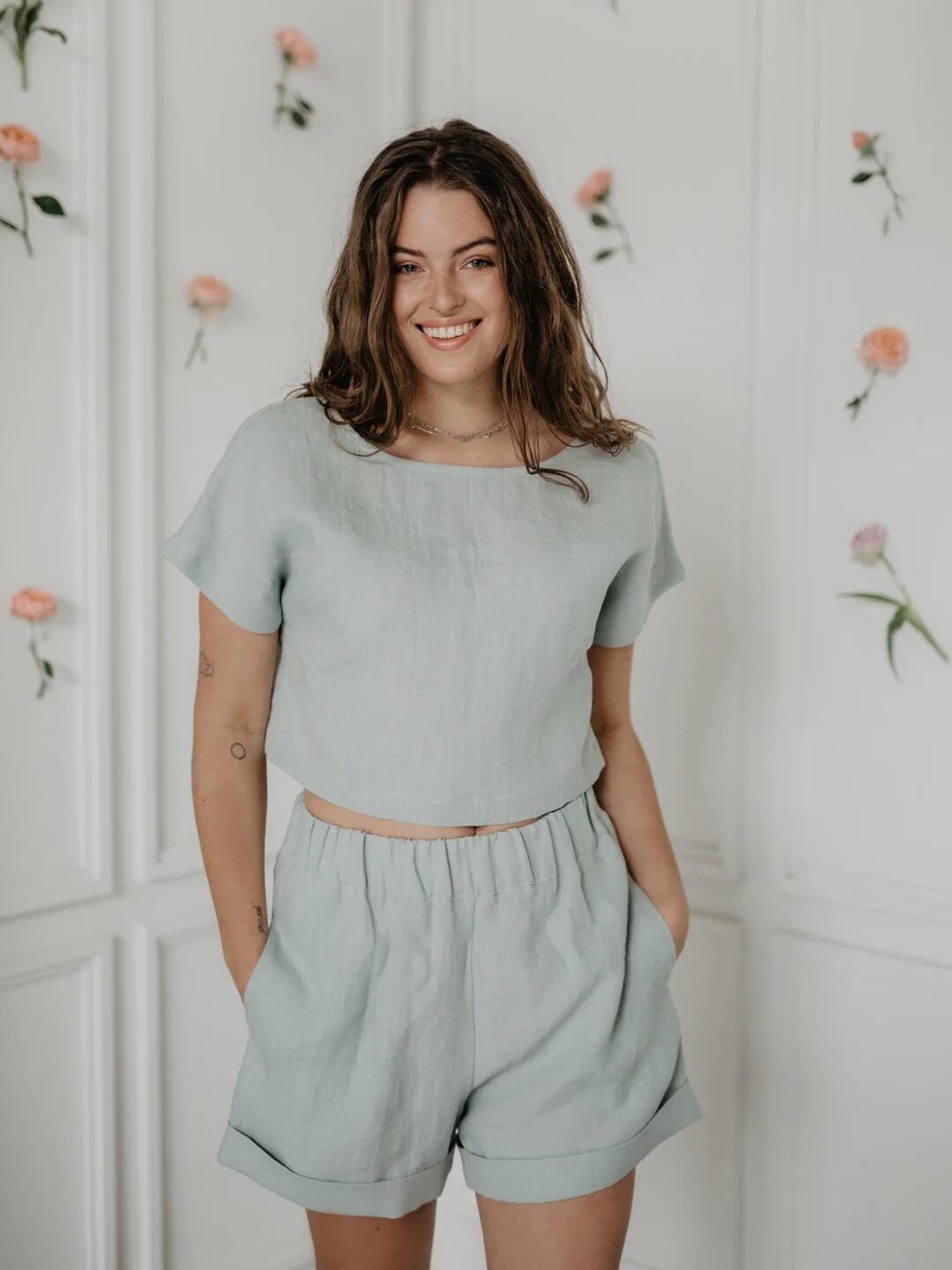 A person with long hair stands smiling, wearing a light green outfit of a short-sleeved top and shorts, with a white wall decorated with flowers in the background.