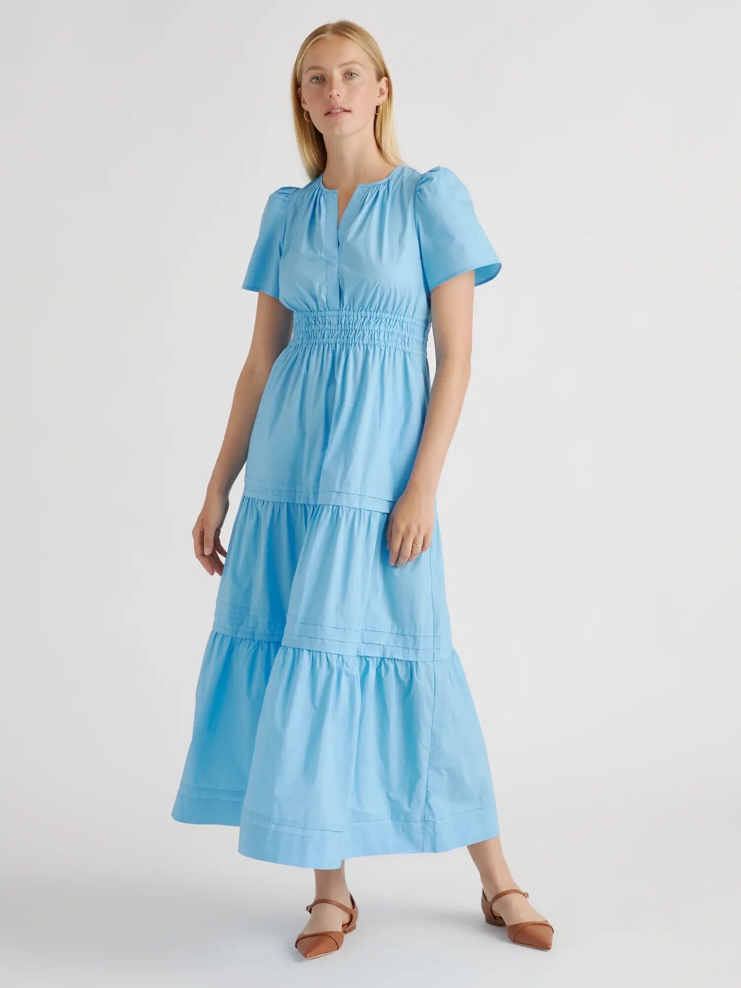 A woman wearing a light blue, short-sleeved, tiered maxi dress stands against a white background. She is also wearing brown flat shoes.