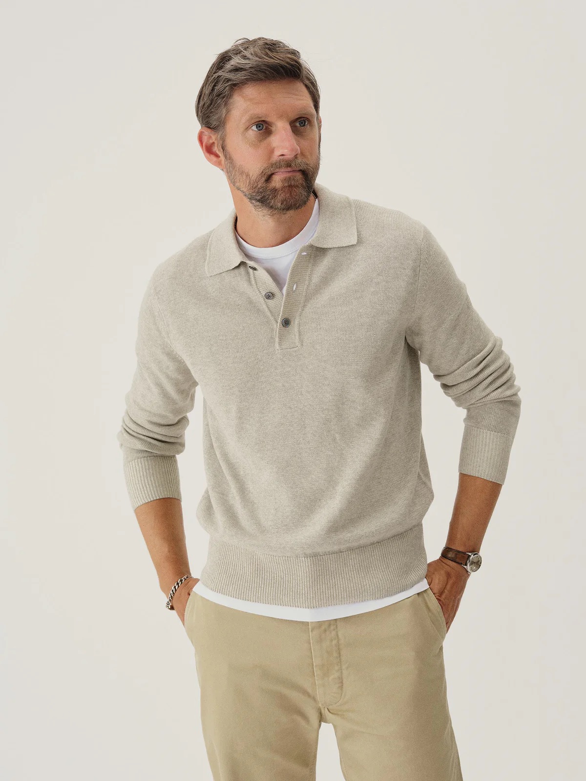 A man with short brown hair and a beard wearing a beige long-sleeve polo sweater and khaki pants stands against a plain background.