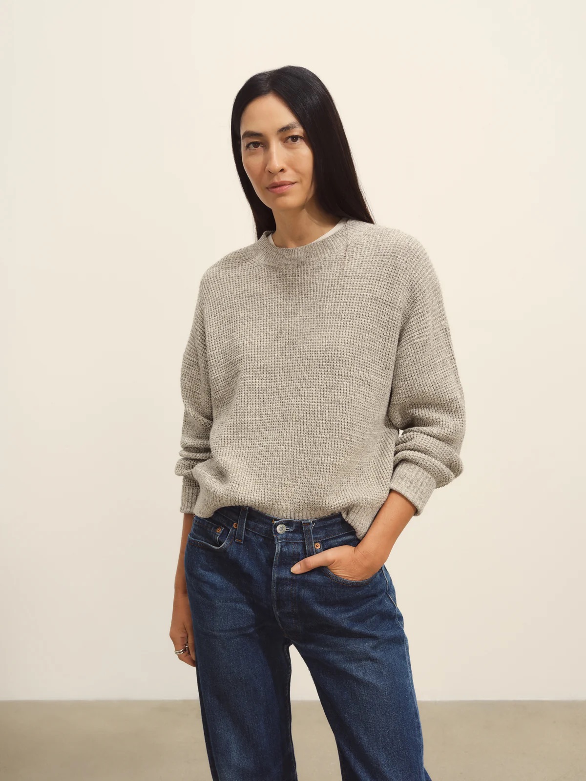 A woman with long dark hair is wearing a loose grey sweater and blue jeans. She is standing against a plain background, with one hand in her pocket.