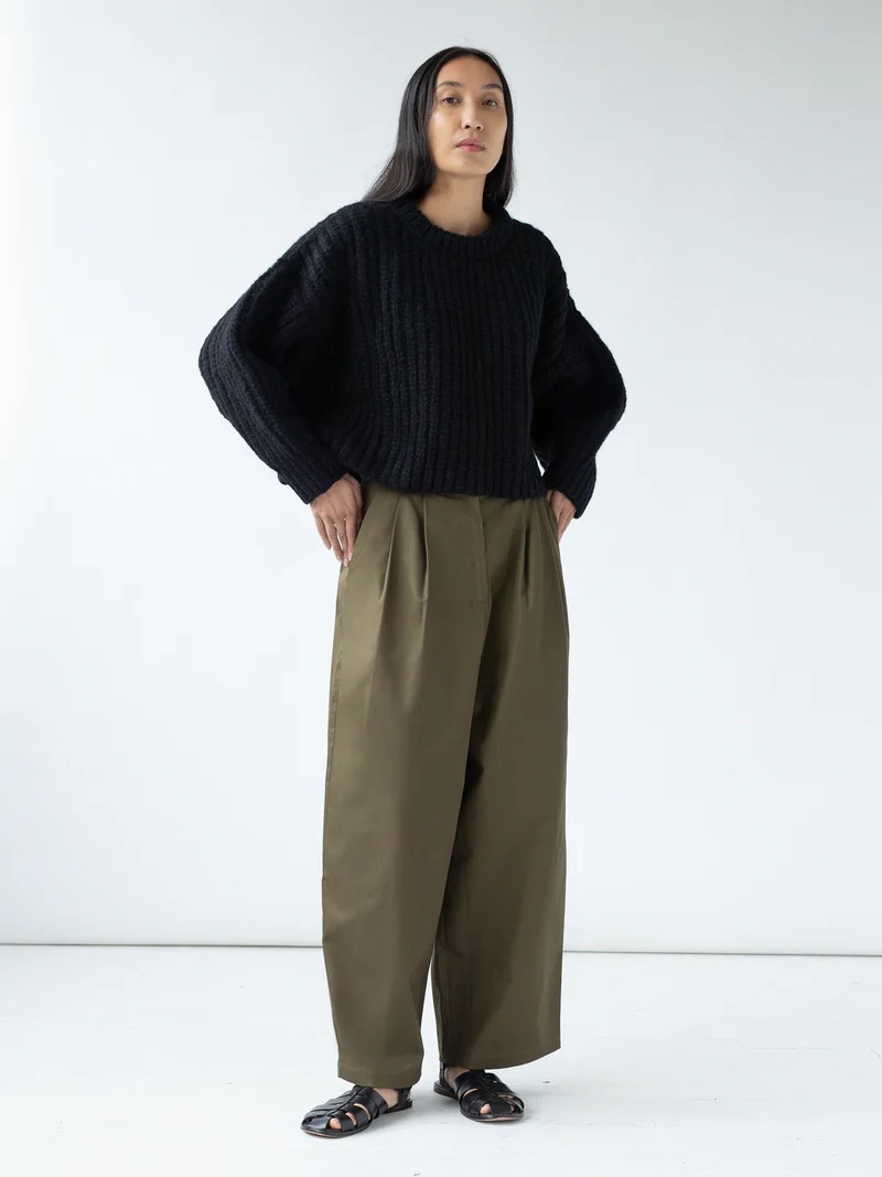 A person with long dark hair stands against a plain background, wearing a black knit sweater, olive green wide-leg trousers, and black sandals.