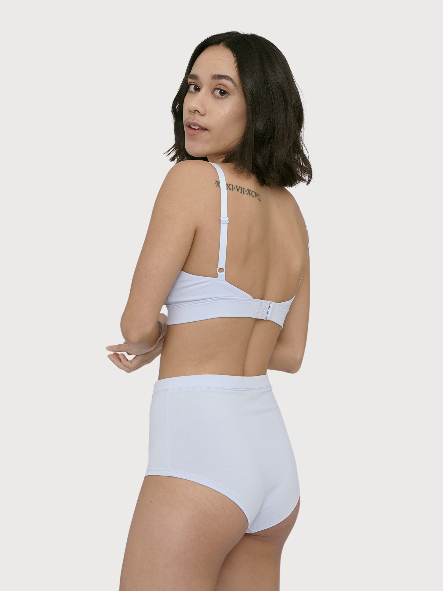 A woman with shoulder-length dark hair poses in white underwear, showing her back and looking over her shoulder. She has a tattoo above her right shoulder blade.