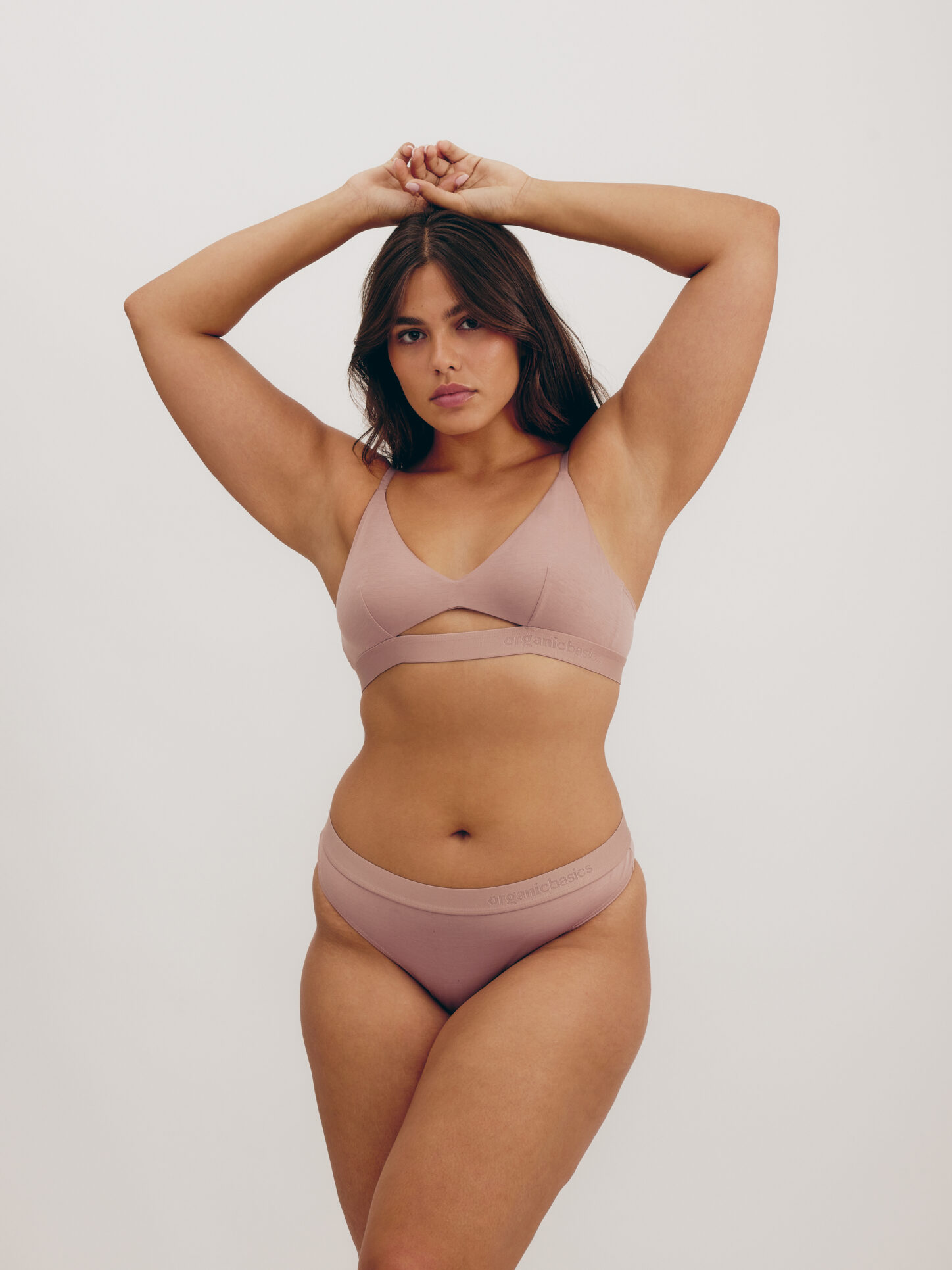 A woman poses with arms raised, wearing a mauve-colored bralette and matching underwear against a plain background.