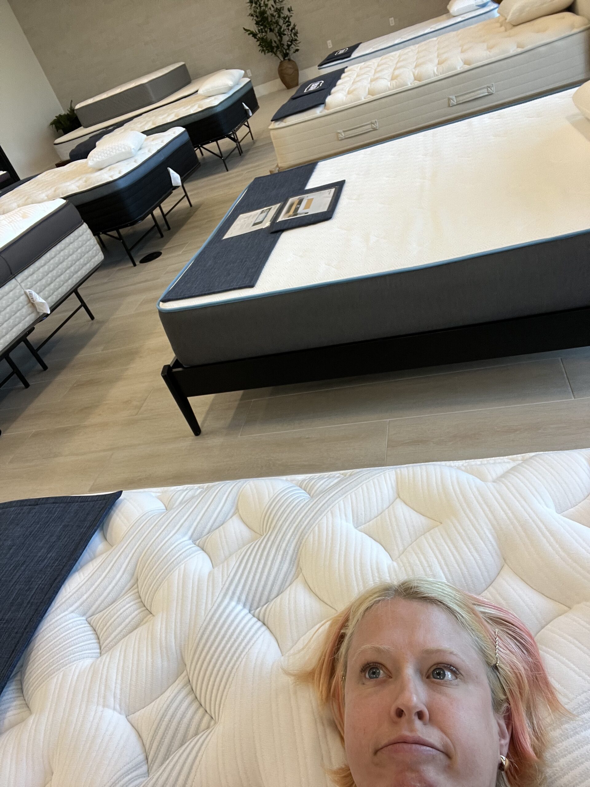 Person lying on a mattress in a showroom filled with neatly arranged mattresses on display.