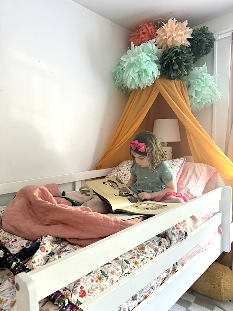 A young child sits in a bed with floral bedding, reading a book. The bed is decorated with a canopy of pastel-colored pom-poms and draped in yellow fabric.