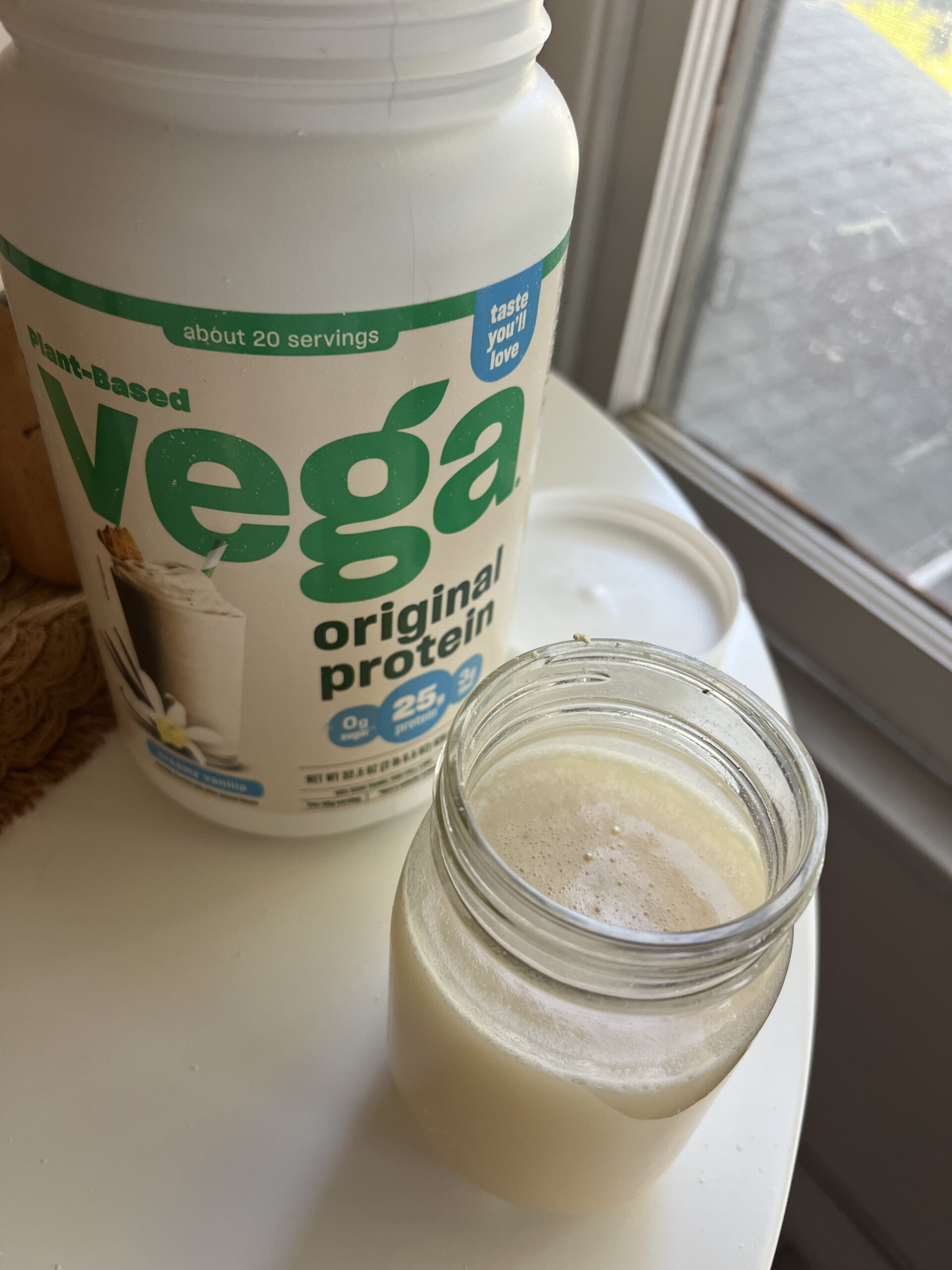 A jar filled with a creamy liquid sits next to an open container of Vega original protein powder on a table.