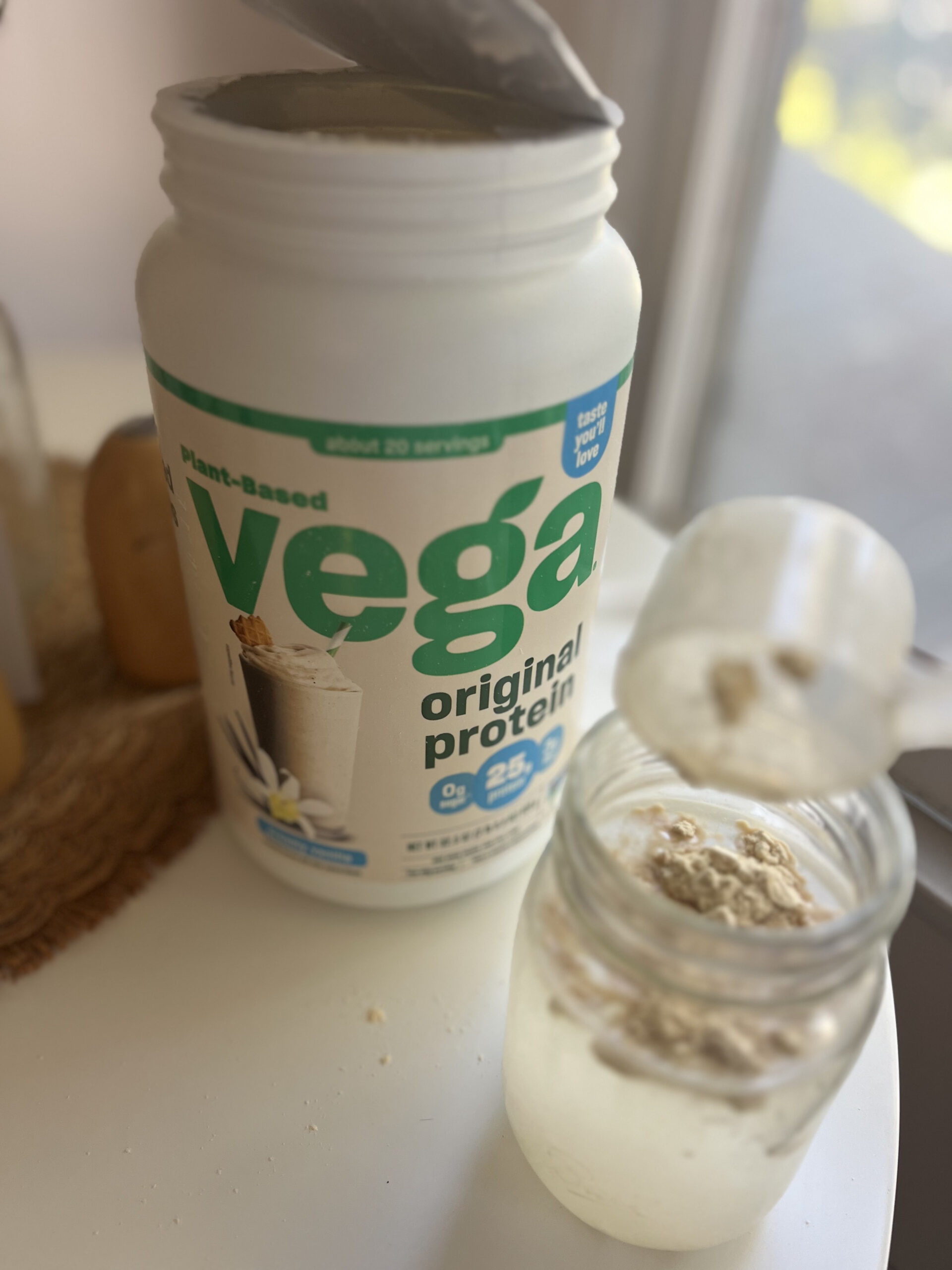 A container of Vega Original Protein powder next to a glass with some powder inside on a white surface.