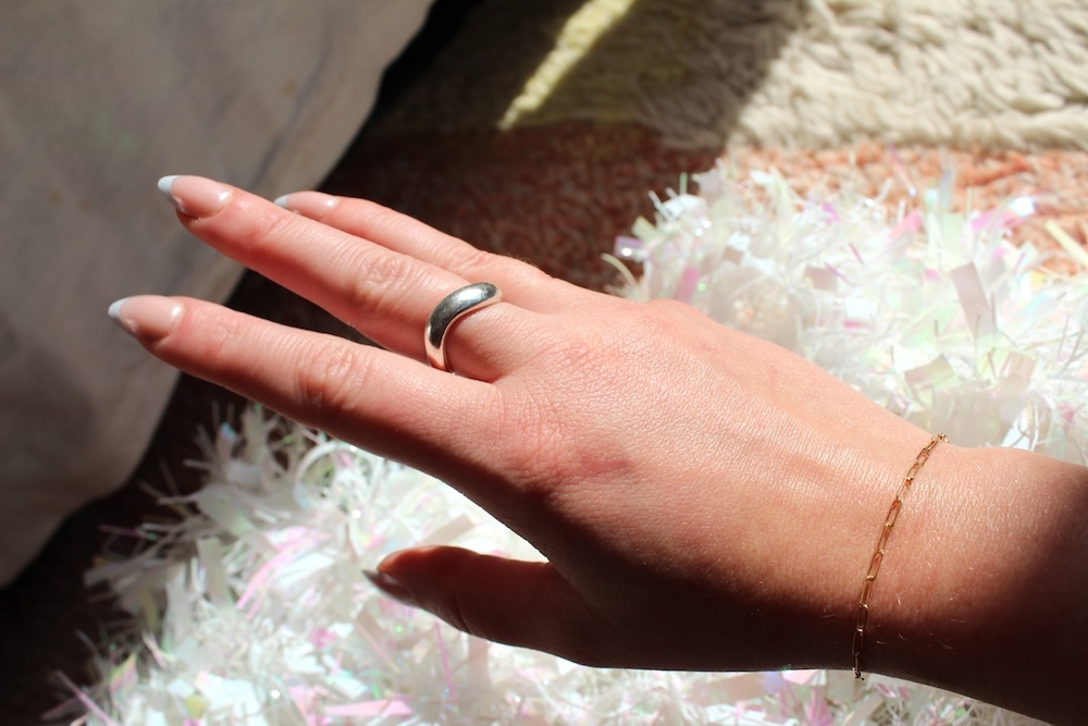 A hand with a silver ring and gold bracelet rests on a surface with white, iridescent material in bright sunlight.