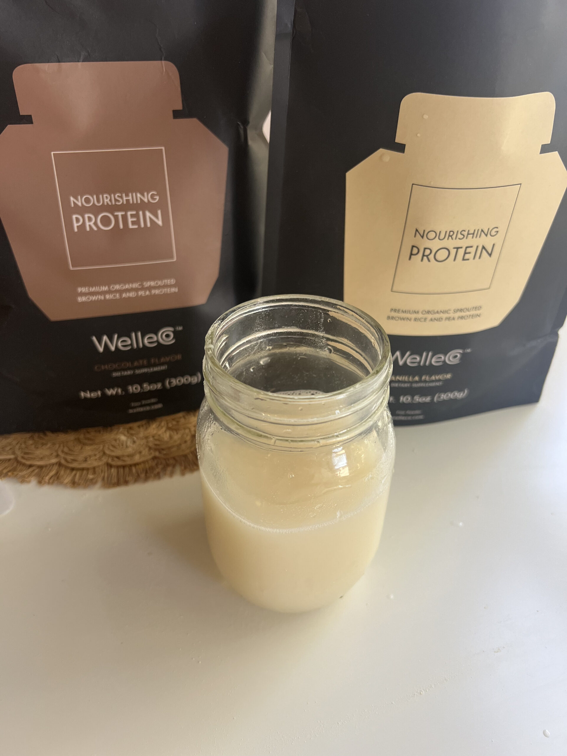 Jar filled with a light-colored drink in front of two pouches labeled "Nourishing Protein" by Welle, one chocolate and one vanilla flavor.