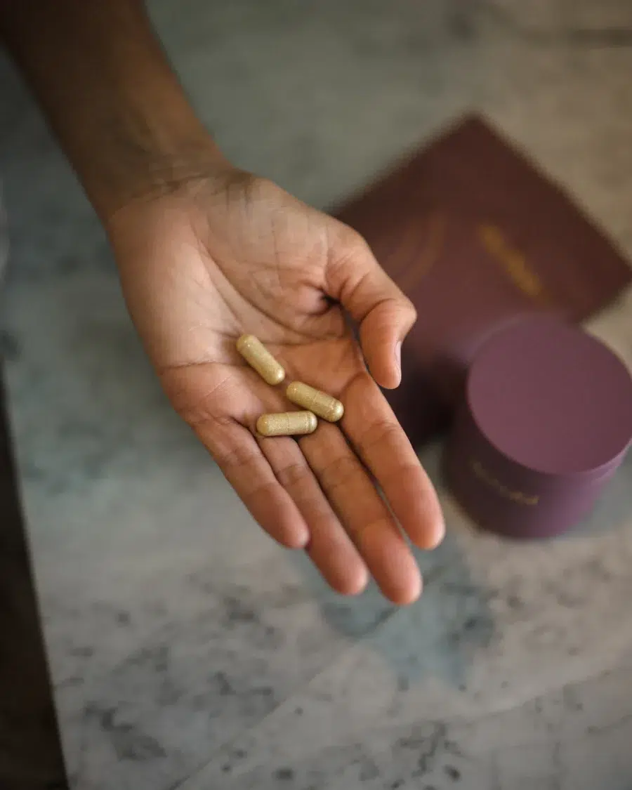 A hand holding three capsule-shaped prenatal vitamins with a blurred background showing a purple container and a similar colored pouch.