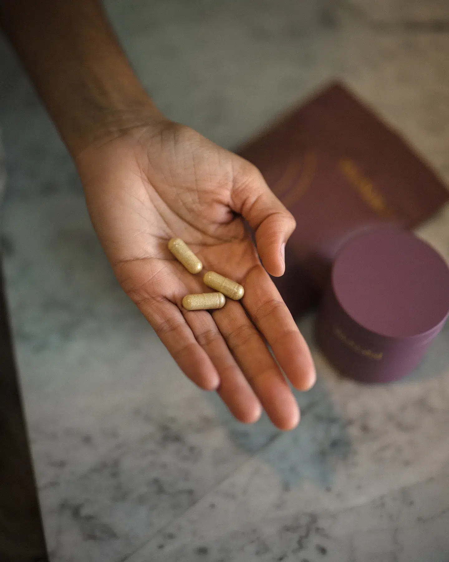 A hand holding three capsule-shaped prenatal vitamins with a blurred background showing a purple container and a similar colored pouch.