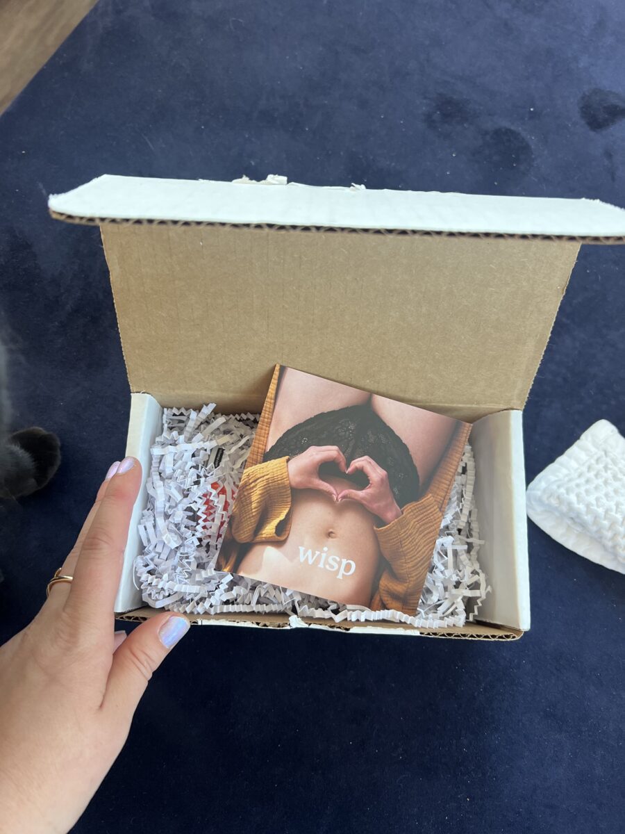 An open cardboard box with white packing material shows a hand holding a brown card with the word “wisp” and a photo of a person forming a heart shape with their hands over their abdomen.