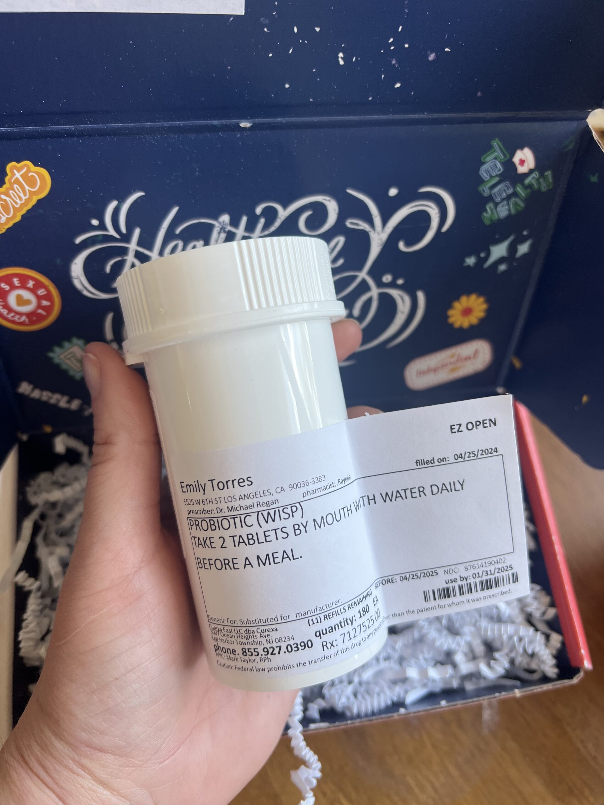 A person holds a white medication bottle with a label that reads "Probiotic (WISP)" with dosage instructions. The bottle is in front of an open box containing decorative stickers.