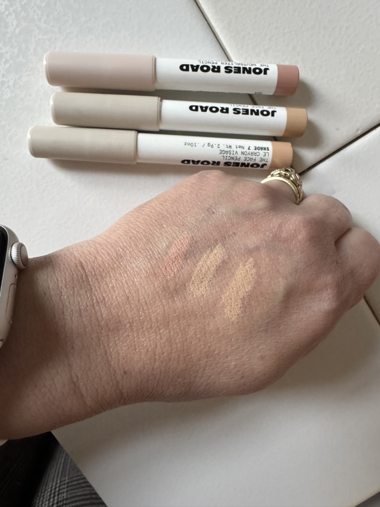 Three makeup sticks labeled "Jones Road" sit elegantly on a tiled surface, while a hand showcases swatches of their rich colors. Reminiscent of Ilia's commitment to quality, these shades promise both vibrancy and depth, making them a staple in any beauty routine.