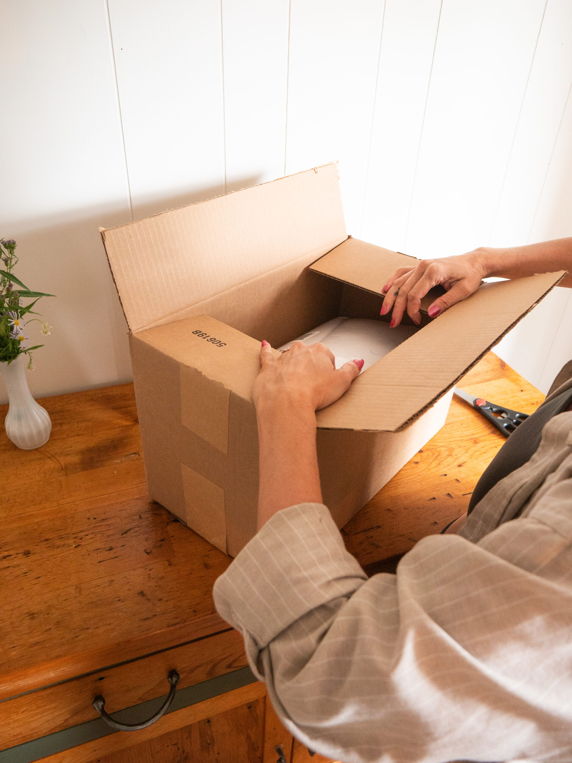 A person opening a cardboard box on a wooden table with a small vase containing greenery in the background.