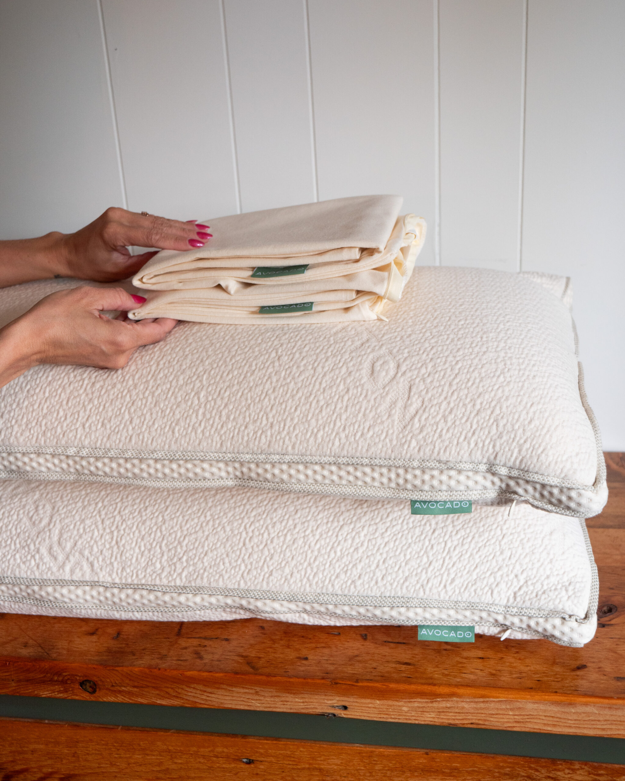 Two hands arranging folded cream-colored pillowcases on top of two stacked white pillows with green tags, placed on a wooden surface against a white background.