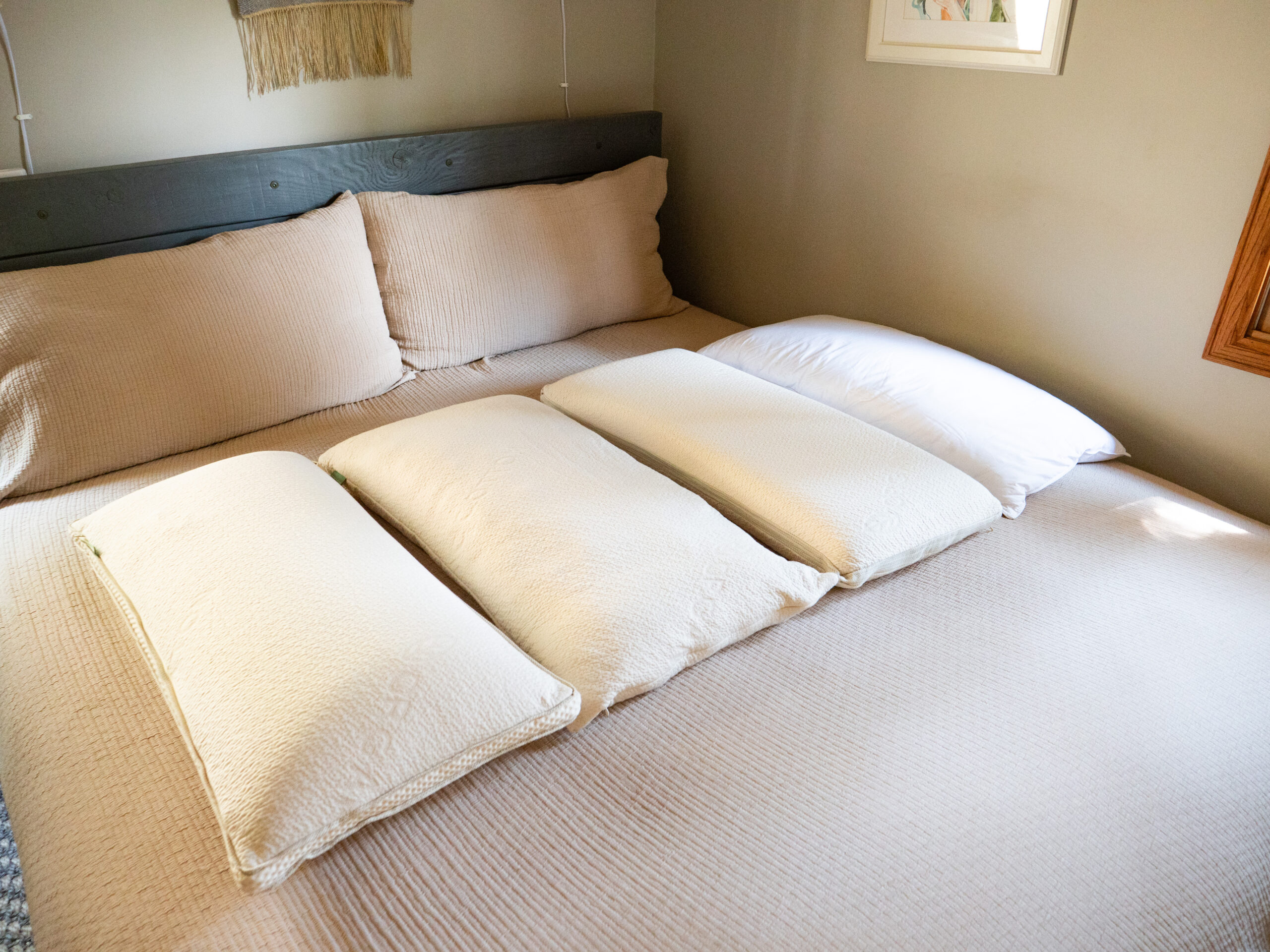 A neatly made bed with a beige bedspread, three beige pillows, and one white pillow arranged in a row. The bed is against a gray wooden headboard in a softly lit room.