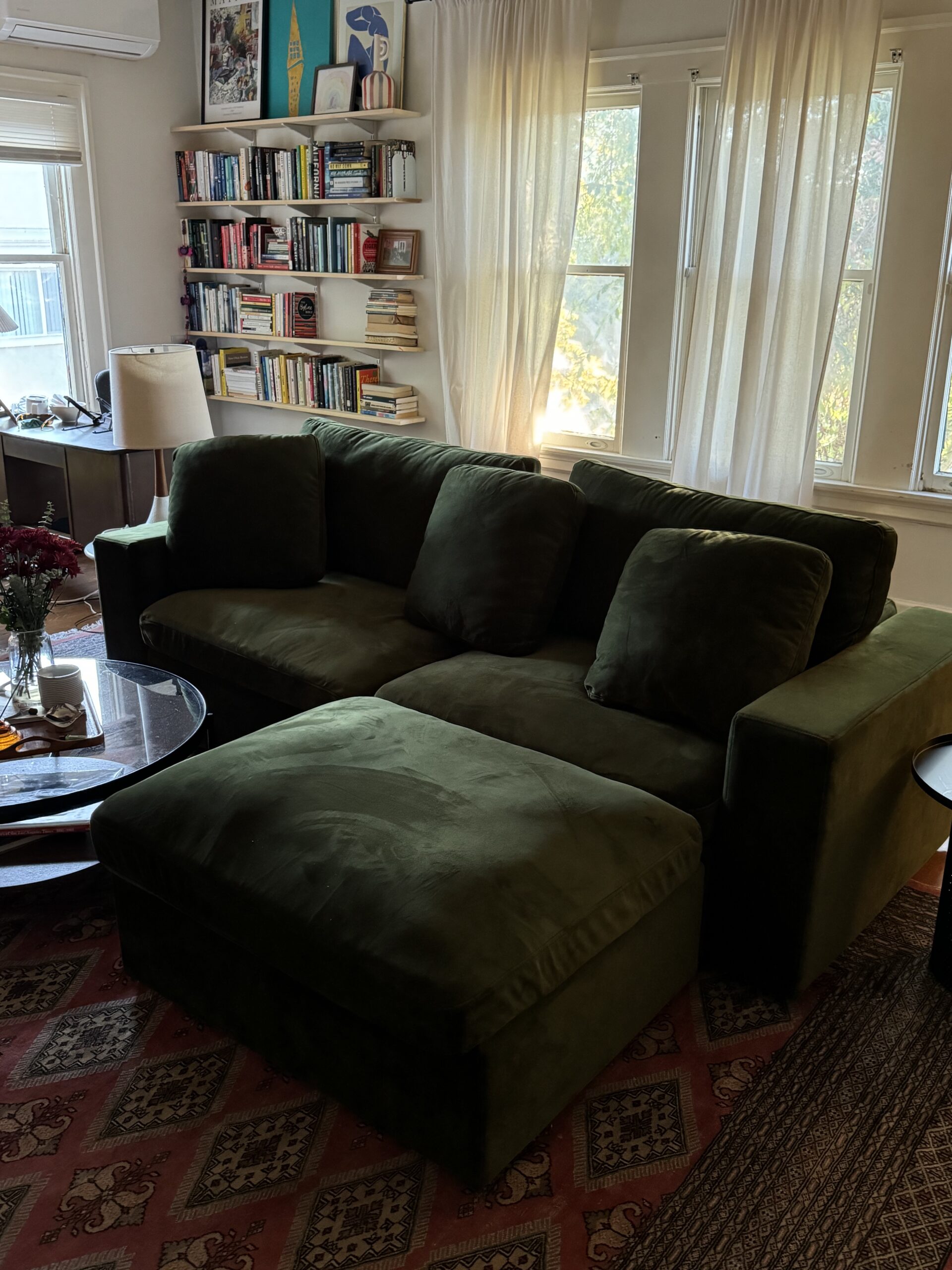 A green sectional sofa in a living room with a circular coffee table, bookshelves, and large windows with curtains.