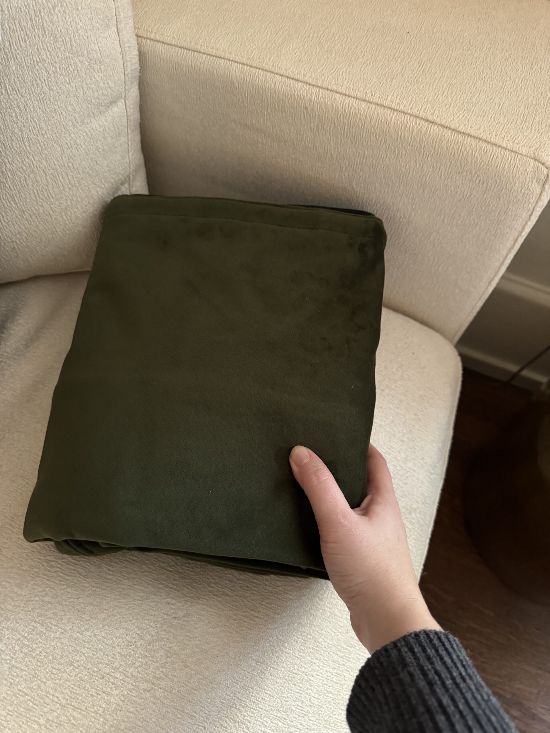 A hand holding a folded, dark green blanket on a light-colored sofa.