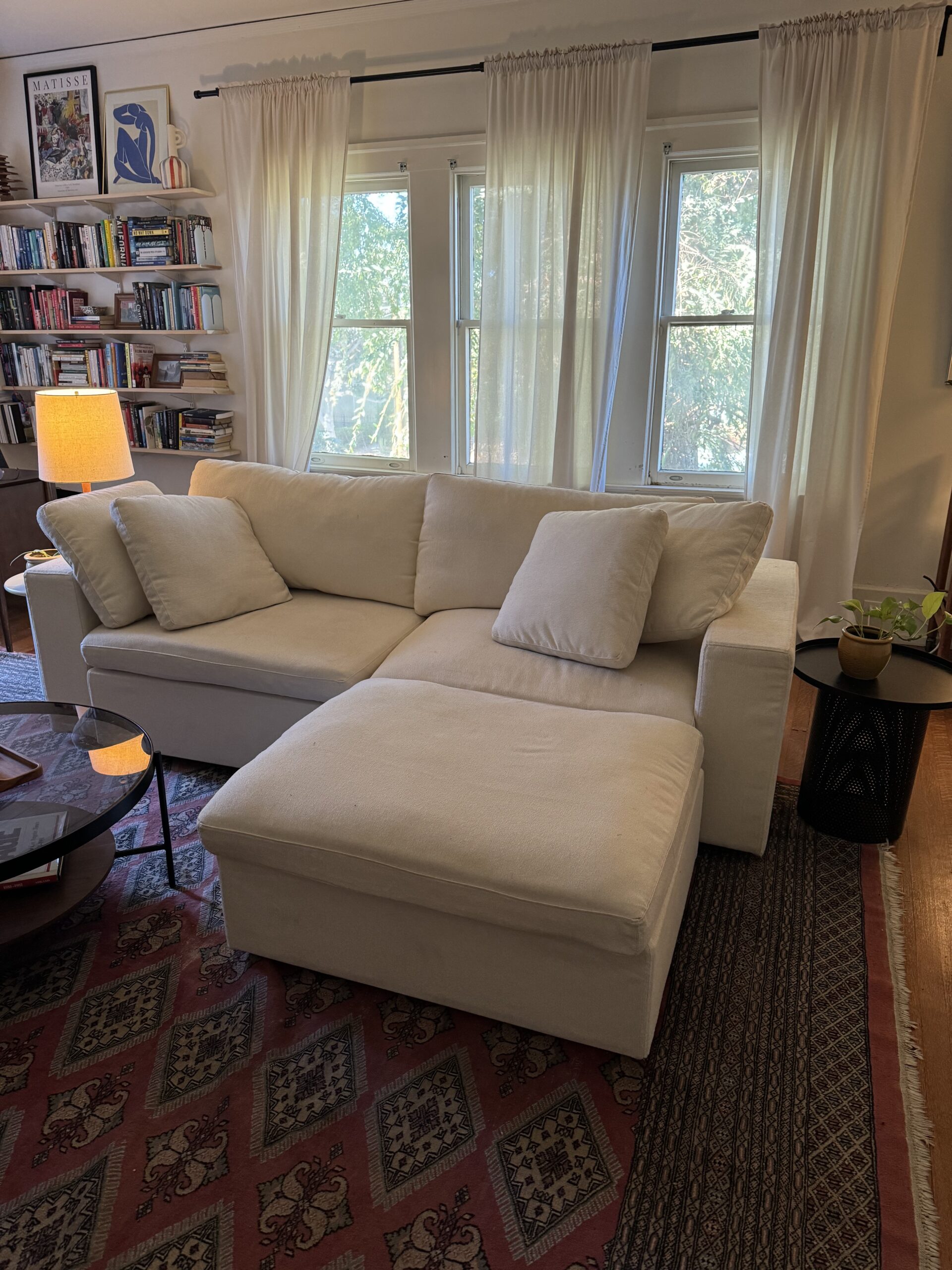 A cozy living room with a white sectional sofa, a patterned rug, a small black side table, and a lit lamp. A bookshelf and windows with sheer curtains are in the background.