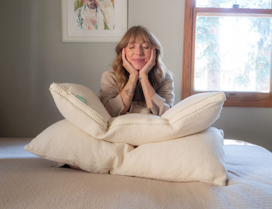 A person with long hair sits on a bed, resting their face in their hands and leaning on a pile of pillows.