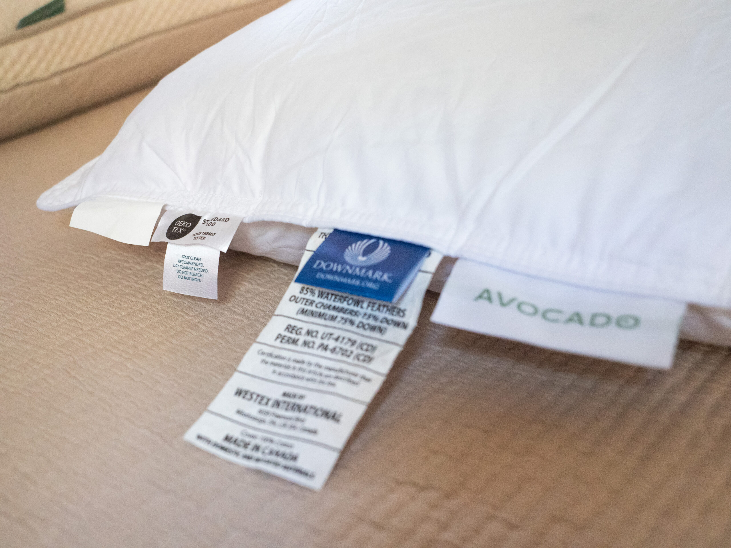 Close-up of a white pillow with several tags showing information about materials and manufacturing, including labels for Downmark, Made in Canada, and Avocado Green Mattress.