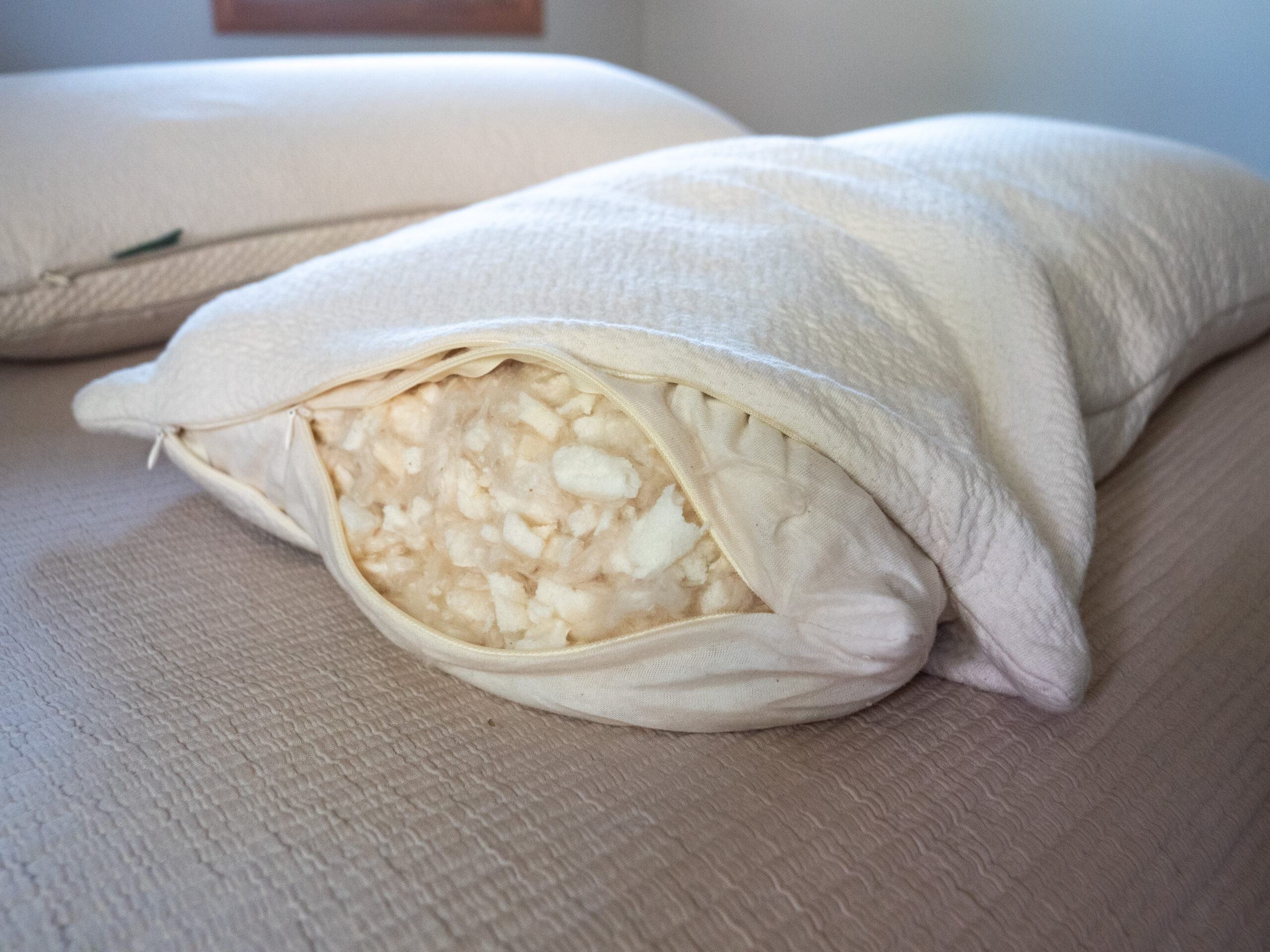 Close-up of a pillow with its cover unzipped, revealing shredded foam filling inside, placed on a bed with another pillow in the background.