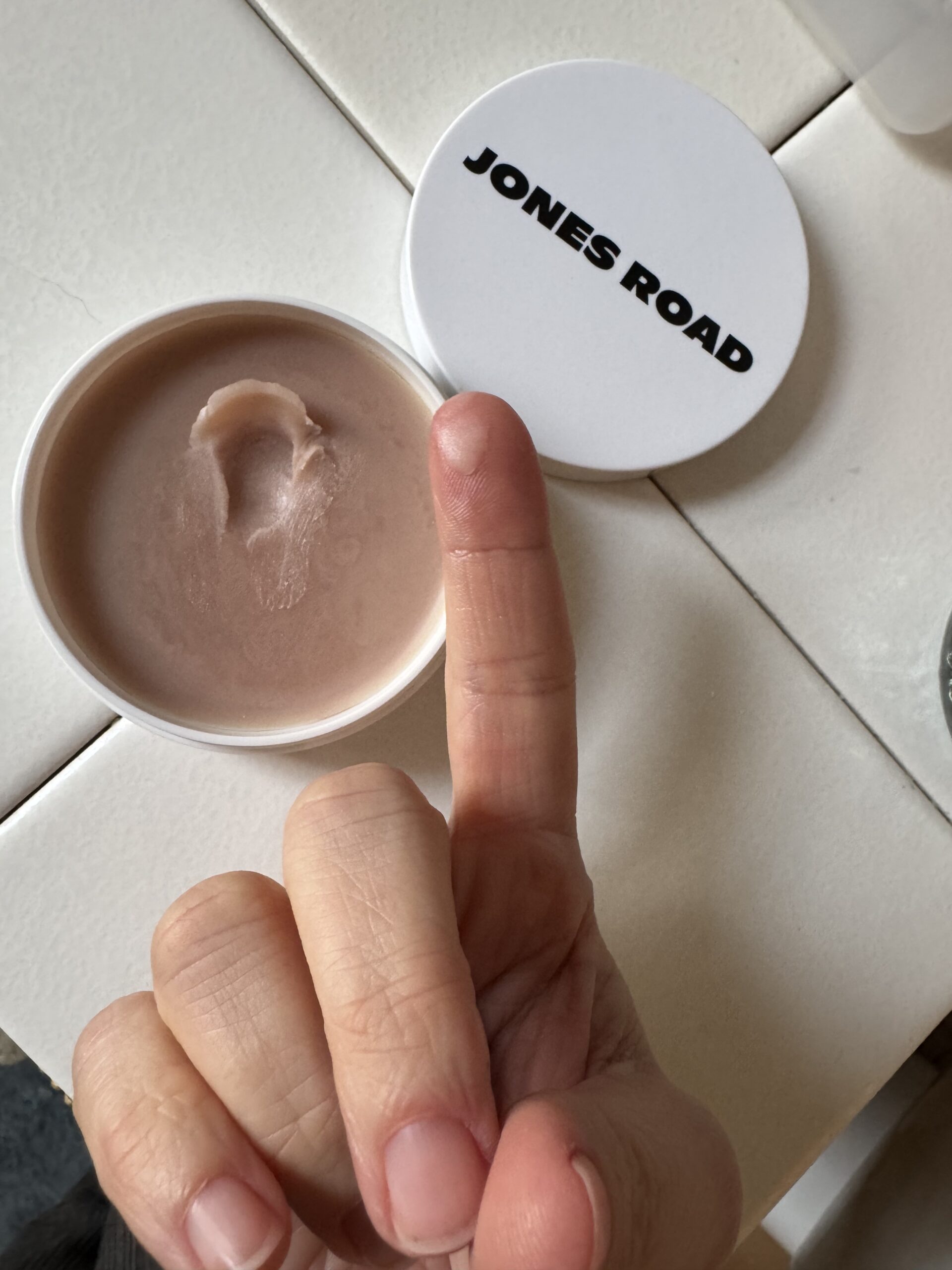 A hand with a finger gently dipped in a beige balm rests in an open container labeled "JONES ROAD" on a pristine white surface