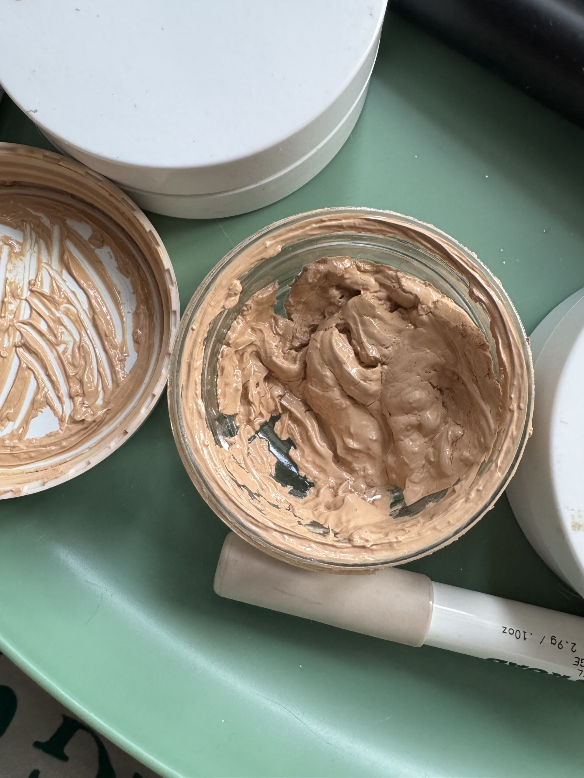 An open jar of What the Foundation from Jones Road sits on a fresh green surface, with some product used and its white lid resting beside it.