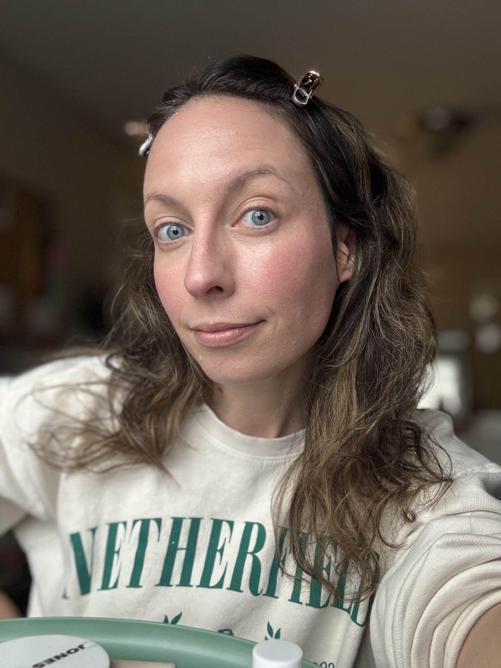 A person with long hair clipped back is wearing a light-colored sweatshirt featuring green text, reminiscent of Ilia's vibrant style.