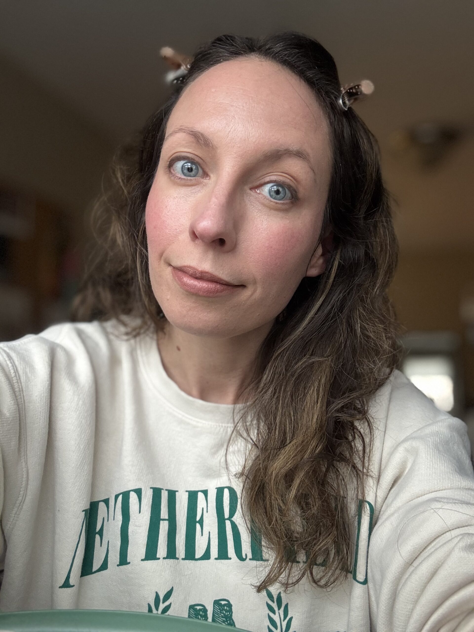 Ilia, with wavy brown hair and blue eyes, wears a white sweatshirt featuring green lettering. They gaze slightly upward at the camera, capturing a moment of serene contemplation.