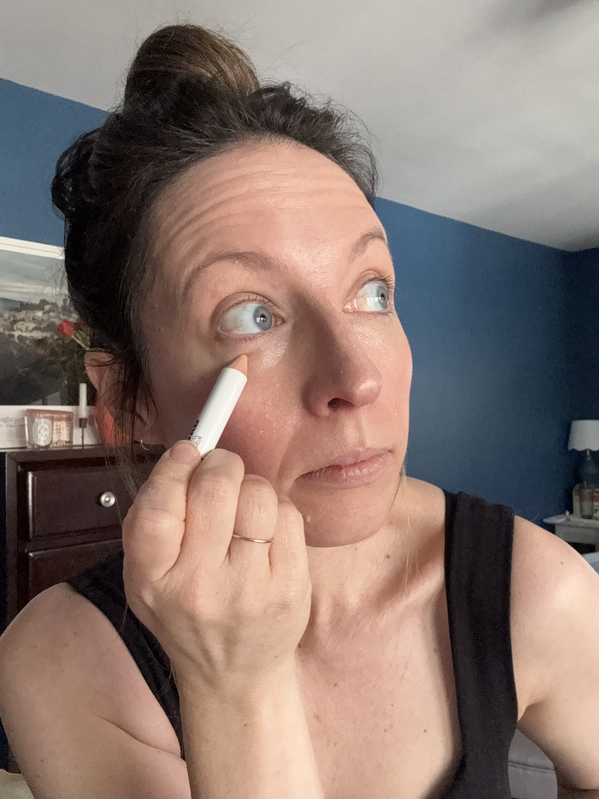 In a cozy bedroom setting, a woman expertly applies makeup under her eye with a white pencil from Ilia, enhancing her natural beauty with every stroke.