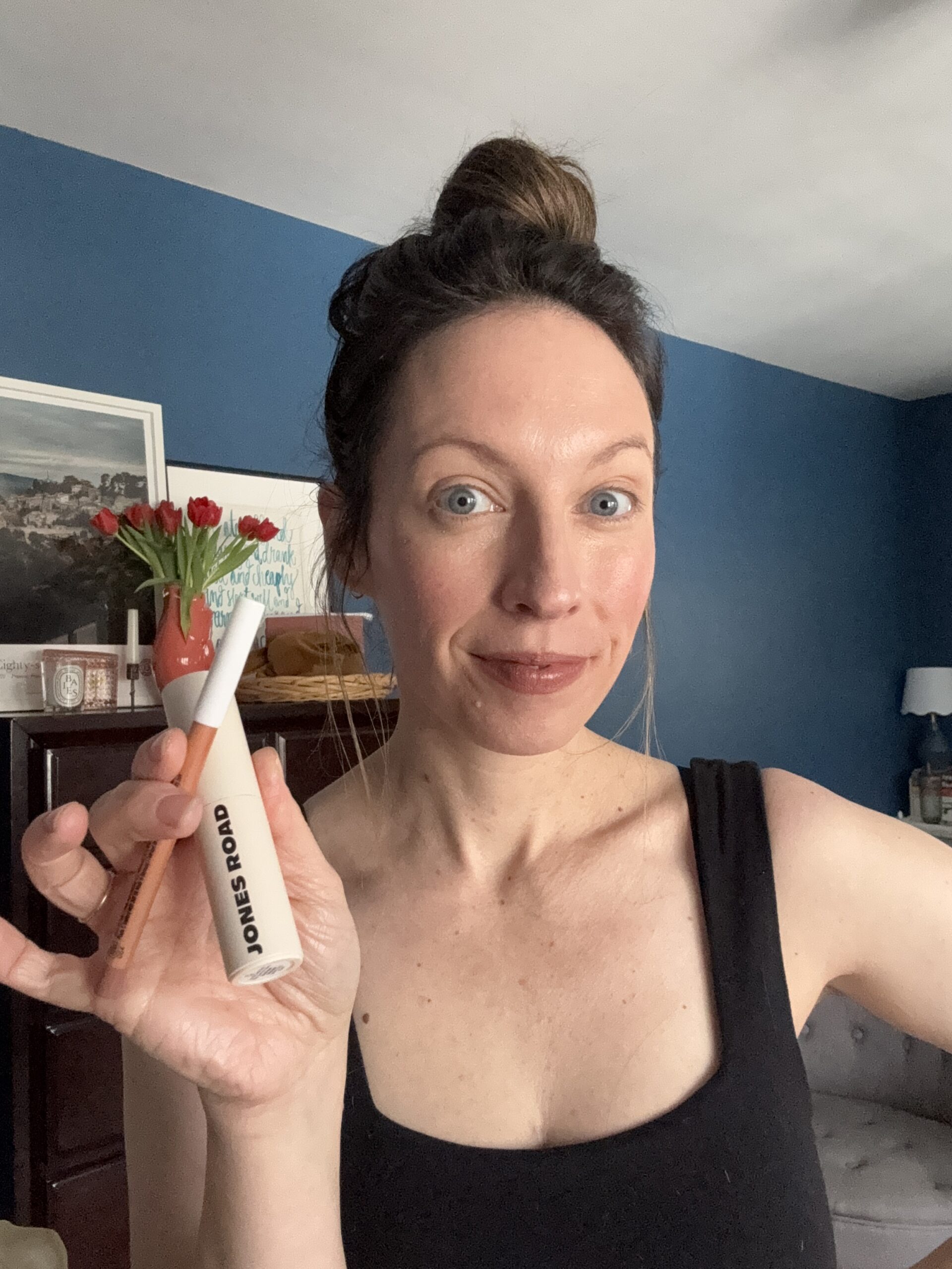 Person in a black top, with hair tied up, gracefully holds a Jones Road makeup product and an Ilia beauty item in one hand, standing in a room adorned with blue walls and a vase of vibrant red flowers.