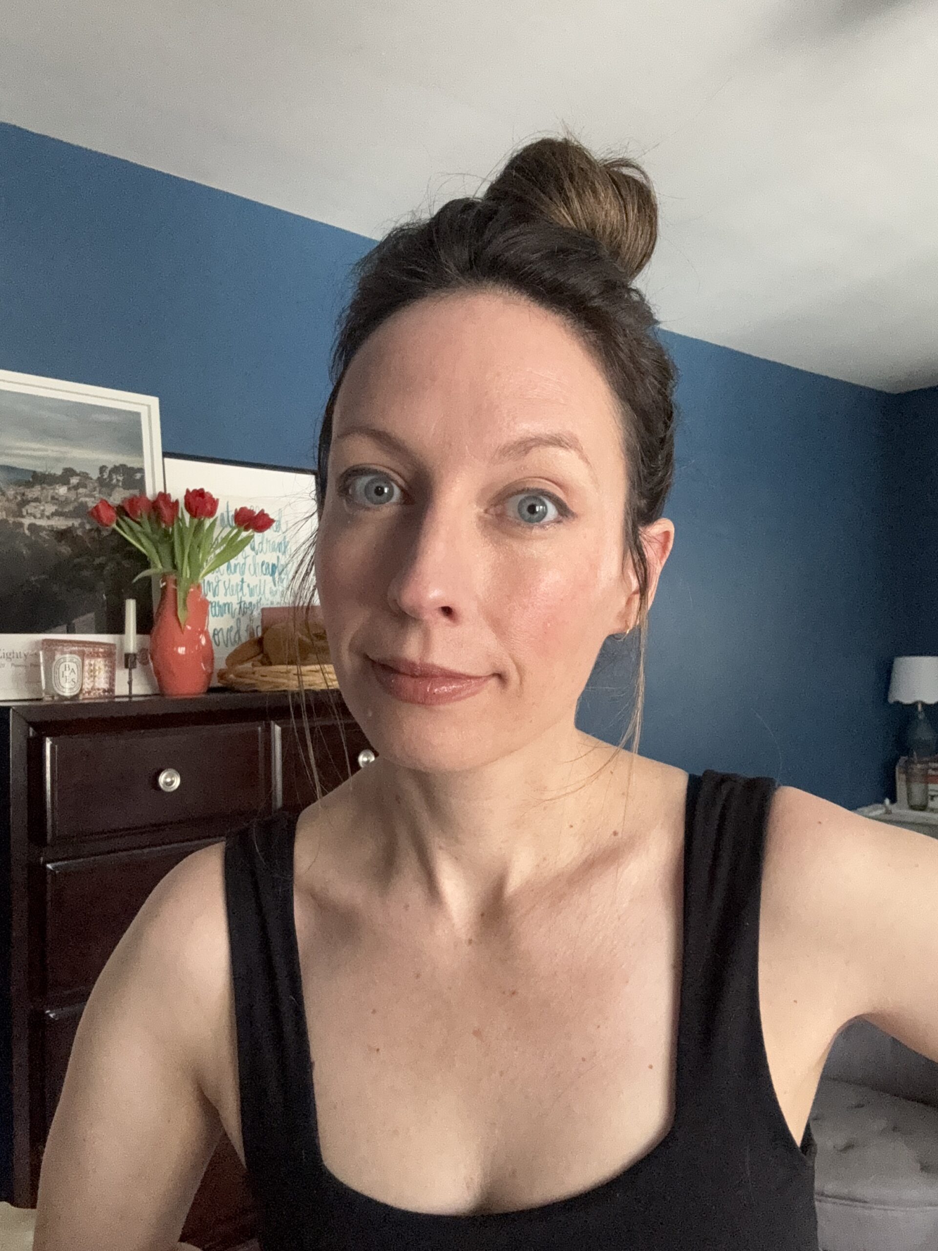 A woman sporting a stylish bun hairstyle and a chic black Ilia top stands gracefully in a blue-walled room, complemented by a dresser, a framed picture, and a vase of fresh tulips.