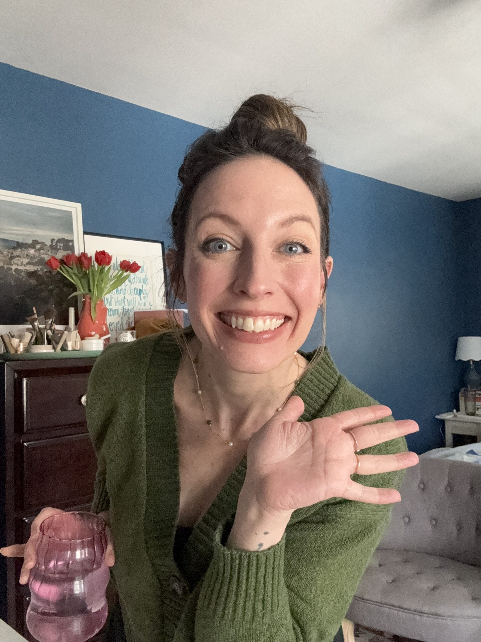 Person smiling and waving at the camera, holding a pink glass. They stand in a room with Ilia-blue walls and a bouquet of red tulips on the furniture in the background.