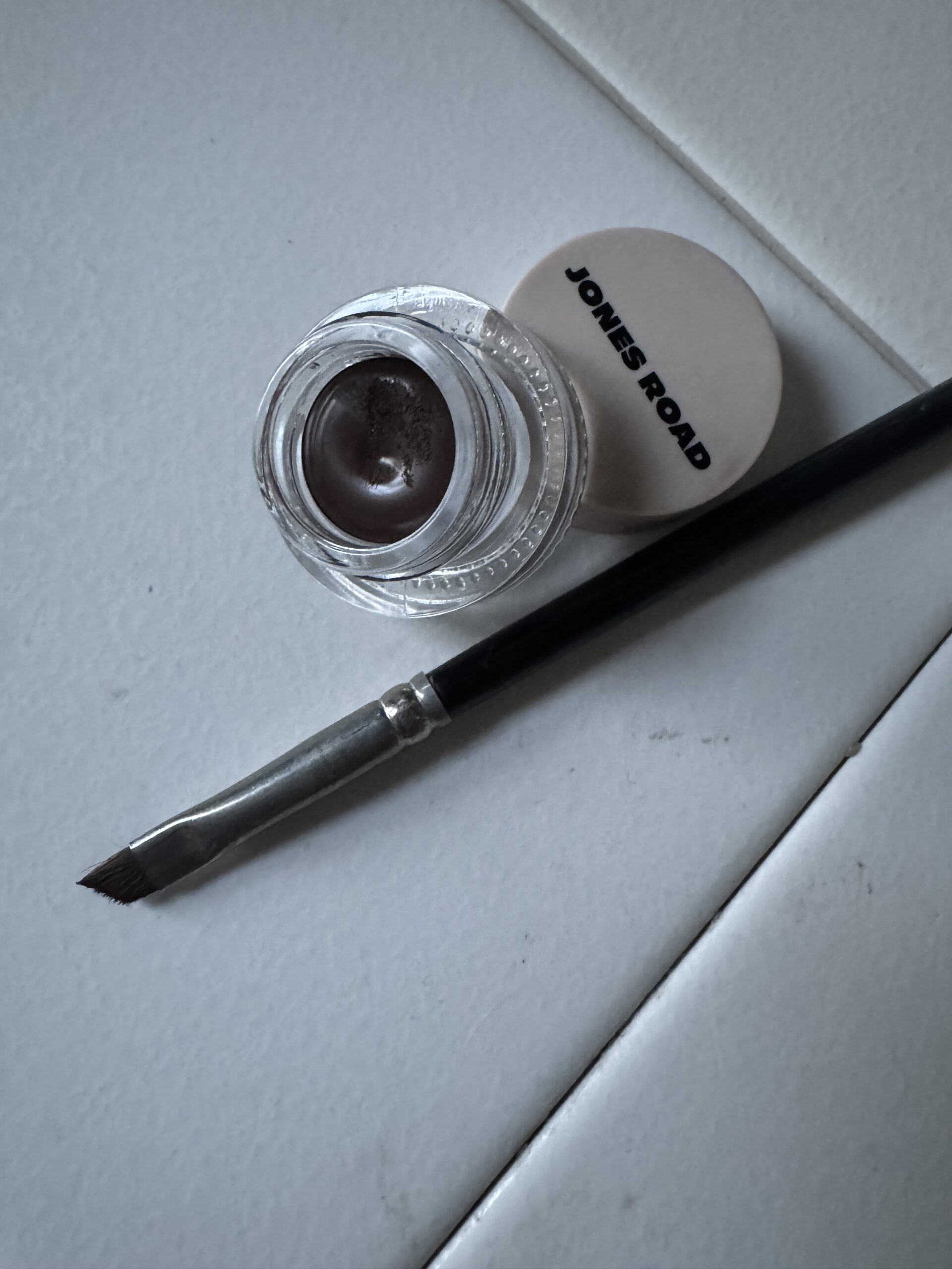 An open jar of makeup with a "Jones Road" lid and an angled brush sits elegantly on the white tile surface, reminiscent of Ilia's clean beauty ethos.