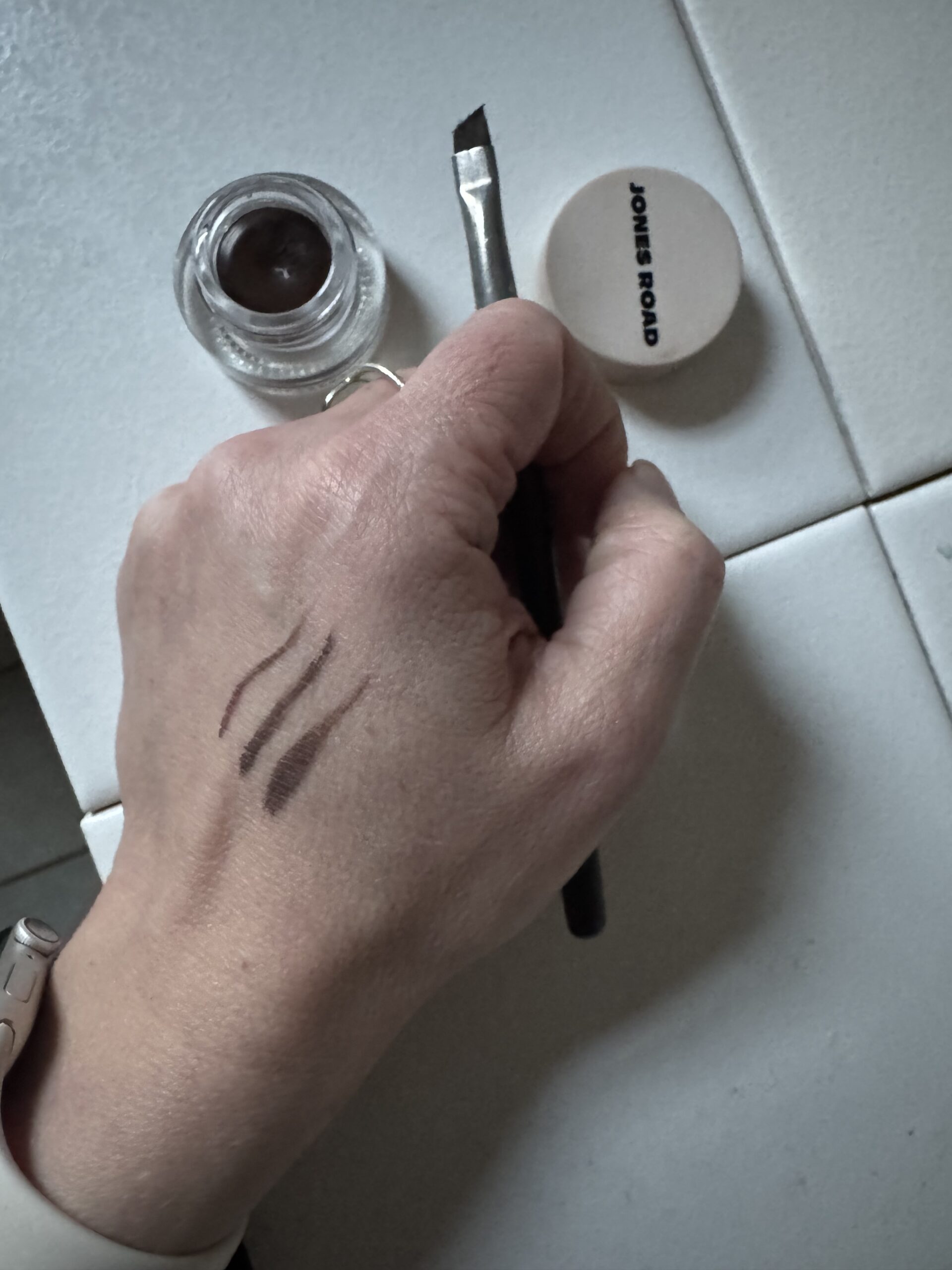 A hand with three brown makeup swatches next to an Ilia brush, holding it gently. Nearby, an open jar of brown gel and a closed lid labeled "Jones Road" rest on the tiled surface.