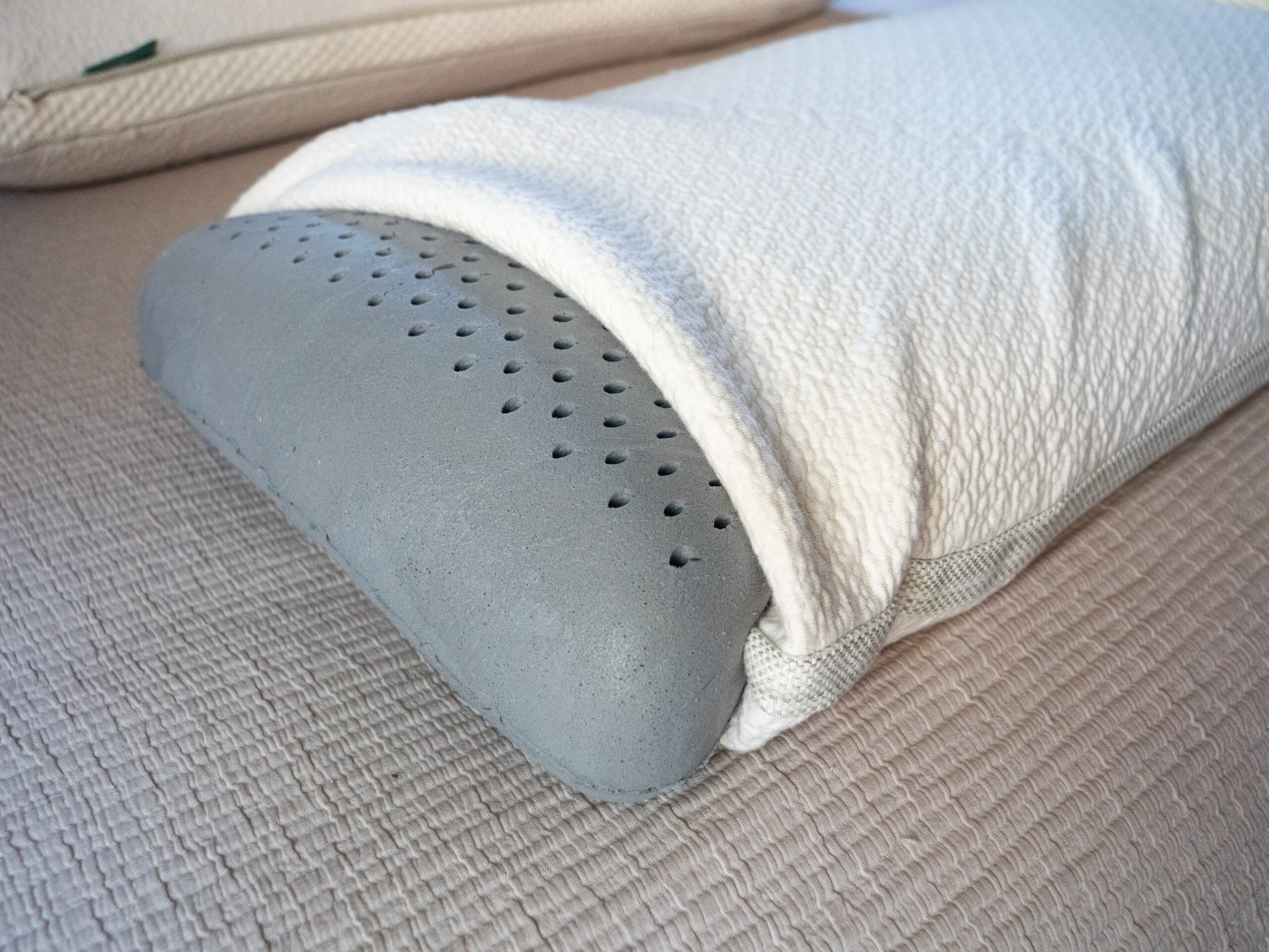 A close-up of a perforated foam pillow partially covered by a textured white cover, placed on a beige, woven fabric surface.