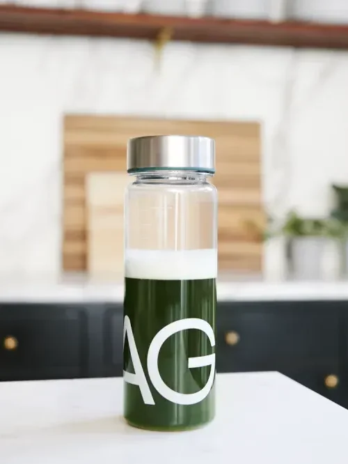 A clear bottle with a silver cap contains a green liquid with a small amount of foam on top. The letters "AG" are printed on the bottle. The bottle is placed on a white surface with a blurred background.