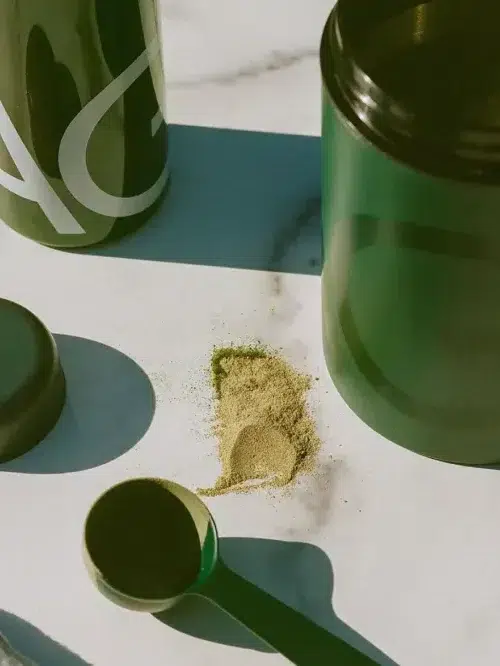 A green powder is spilled on a marble surface next to an open container, a measuring scoop, a lid, and a bottle.